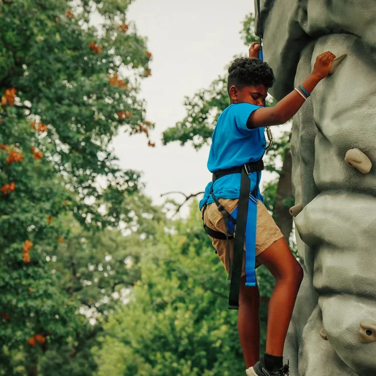 A camper in a harness climbing the outdoor rock wall at camp